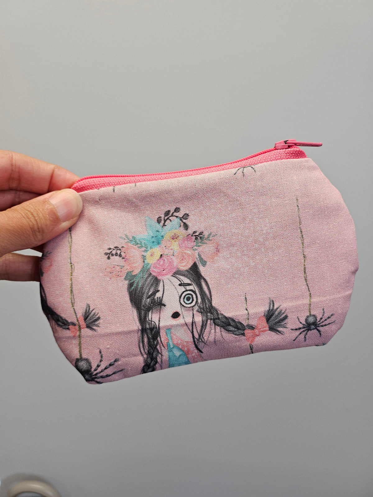 A hand holding a light pink zippered pouch decorated with a gothic girl illustration, featuring a floral crown, black hair, and spider motifs, set against a plain gray background.