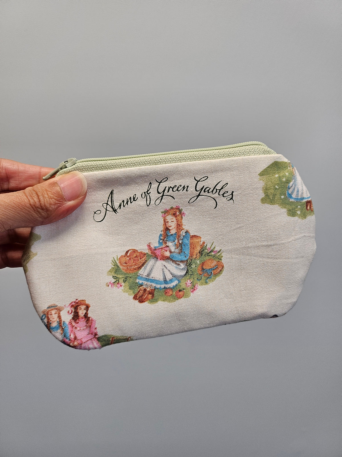 A hand holding a small zippered pouch featuring an Anne of Green Gables illustration and text on a plain gray background.