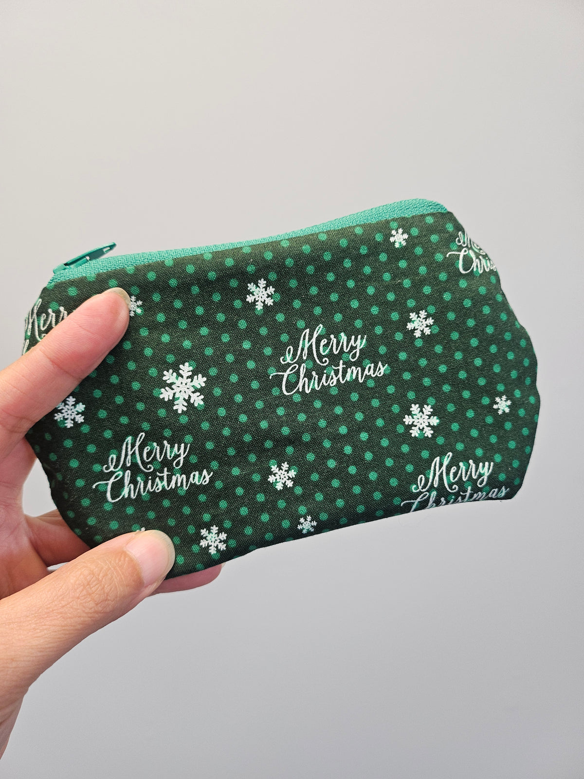 A hand holding a small zippered pouch with a green and white holiday pattern featuring snowflakes and 'Merry Christmas' text, set against a plain light gray background.