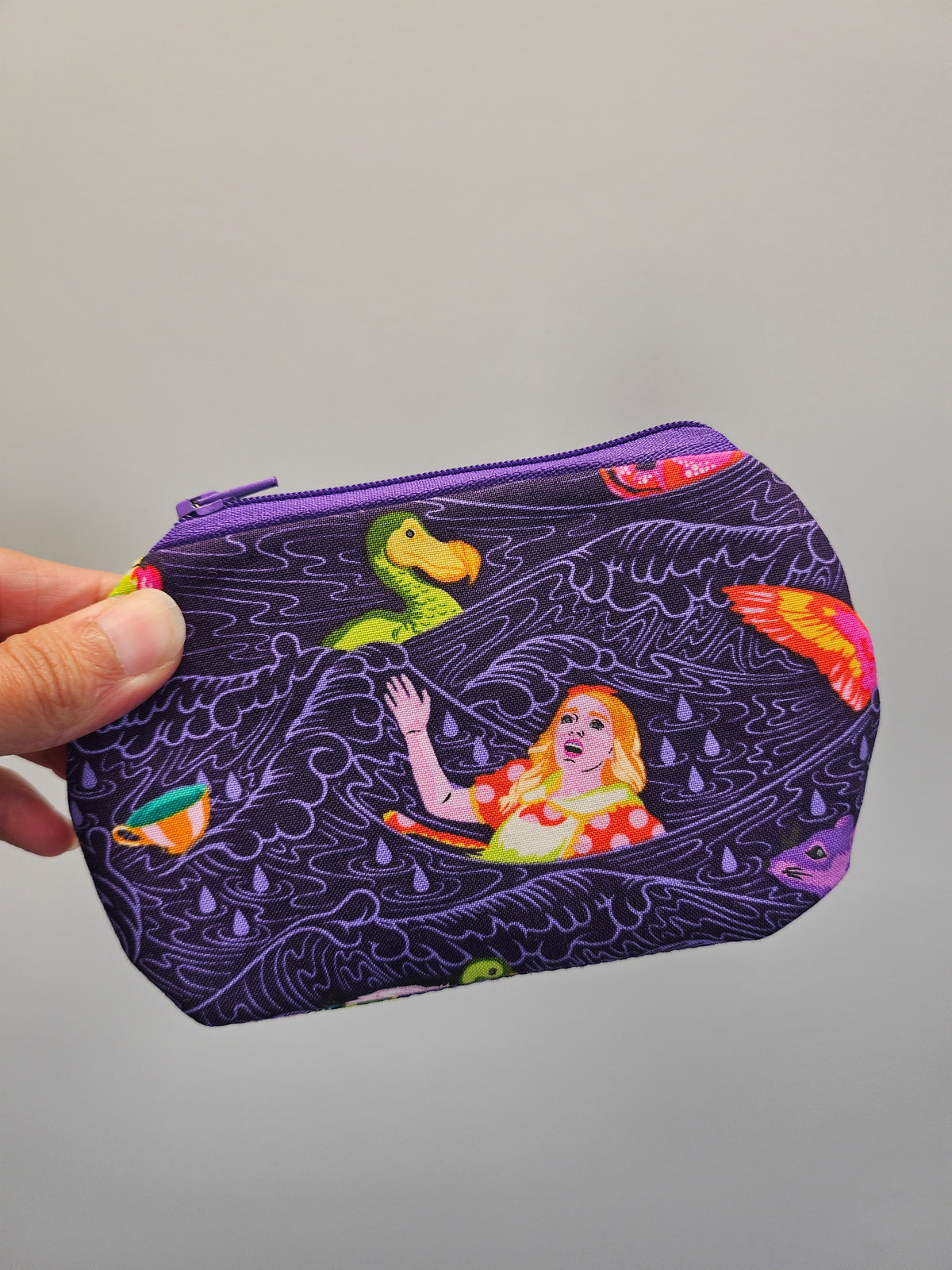 A hand holding a small zippered pouch with a purple background and a whimsical illustration of a woman in a raft with a green duck and colorful fish.