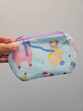 Load image into Gallery viewer, A hand holding a small zippered pouch with a light blue background and a whimsical cartoon print of a pink llama and a yellow cat knitting, featuring a purple zipper.
