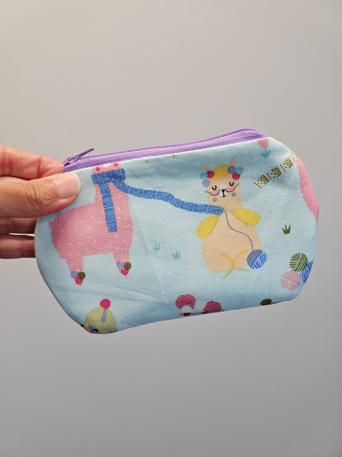 A hand holding a small zippered pouch with a light blue background and a whimsical cartoon print of a pink llama and a yellow cat knitting, featuring a purple zipper.