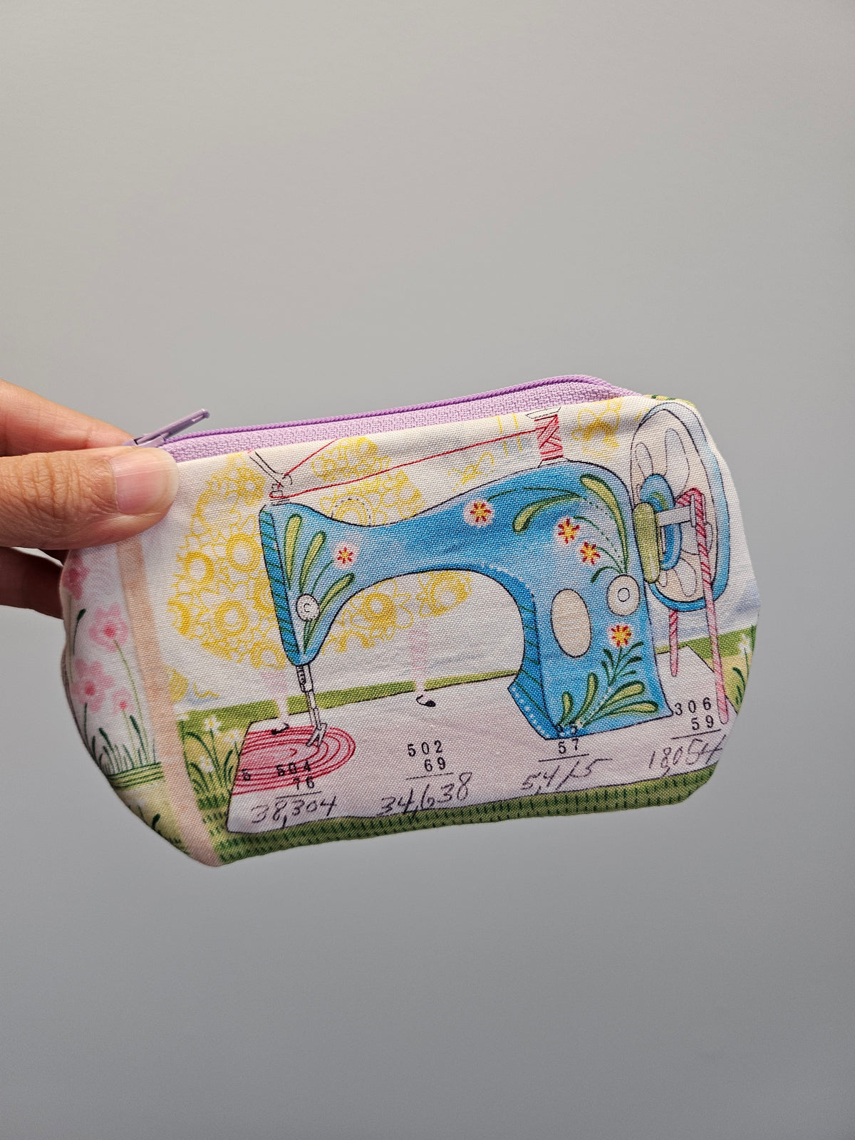 A hand holding a small zippered fabric pouch featuring a colorful printed sewing machine design on a light background.