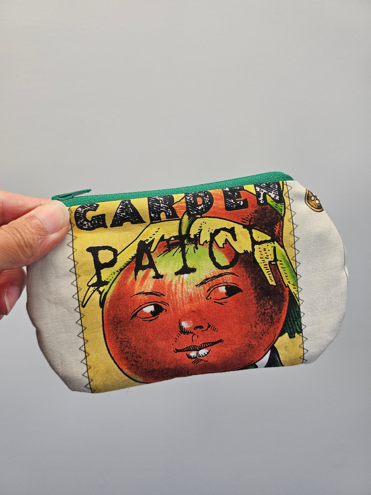 A hand holding a small zippered pouch with a vintage-style printed design featuring a red tomato face and the words 'GARDEN PATCH'. The pouch has a green zipper and a white fabric base.