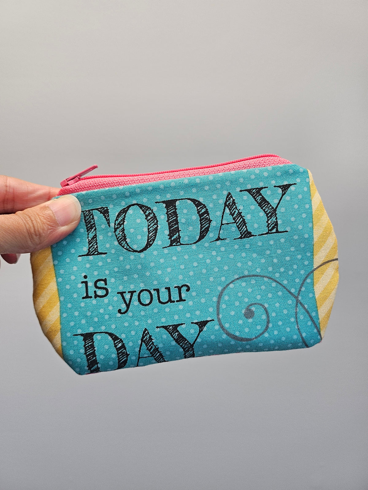 A hand holding a small fabric zip pouch with a turquoise blue polka-dot front panel that reads 'TODAY is your DAY' in black lettering, with yellow-and-white striped fabric on the sides and a pink zipper.