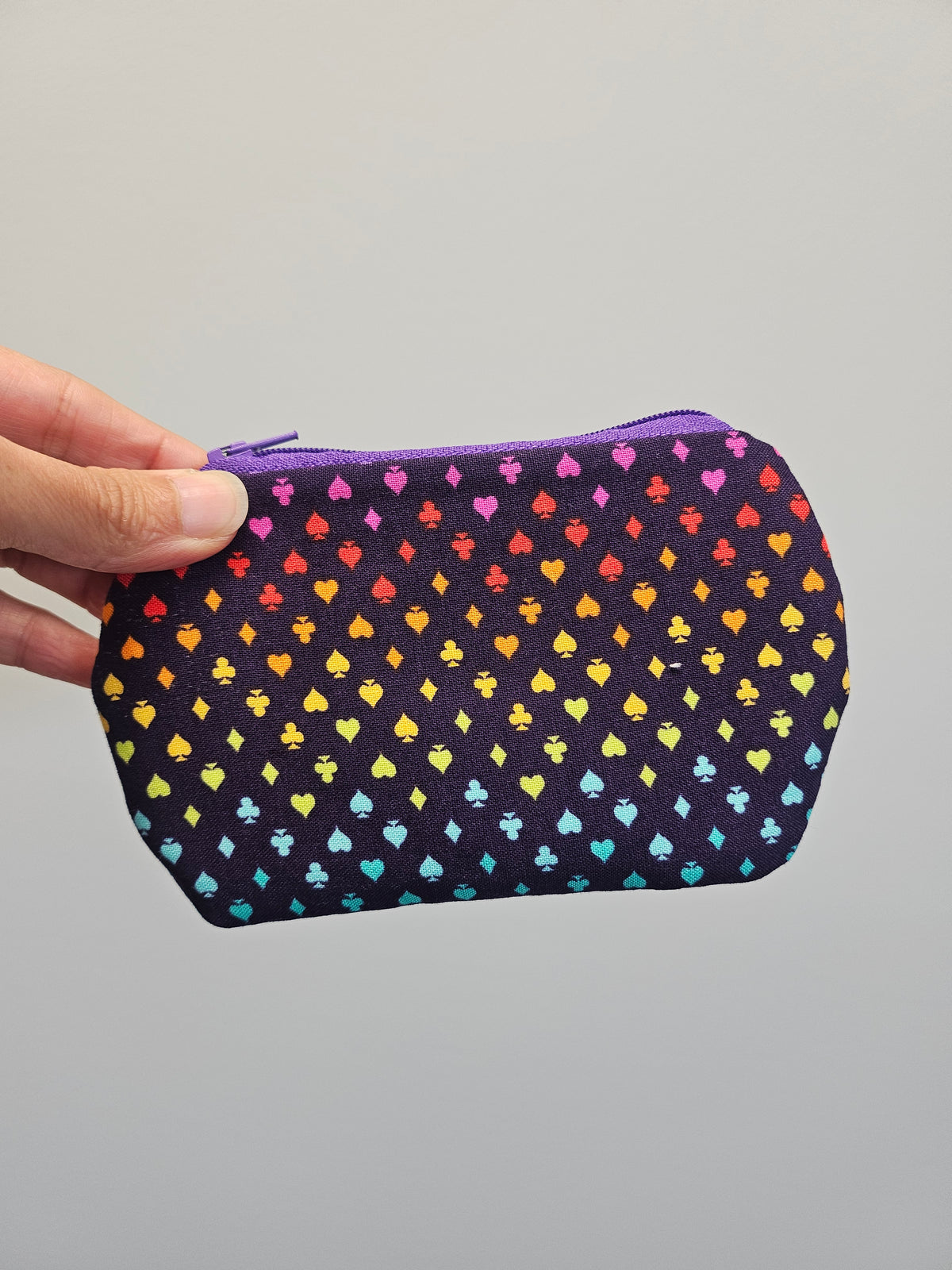 A hand holding a small zippered pouch with a dark purple background and a repeating pattern of small hearts in rainbow colors, against a plain light gray background.