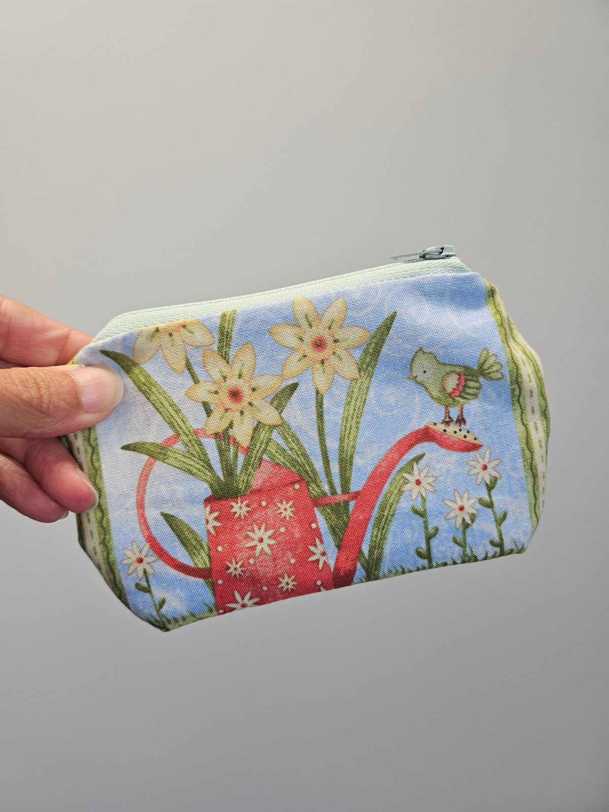 A hand holding a zippered fabric pouch decorated with a whimsical garden print featuring a red watering can, yellow flowers, green leaves, and a small bird on a light gray background.