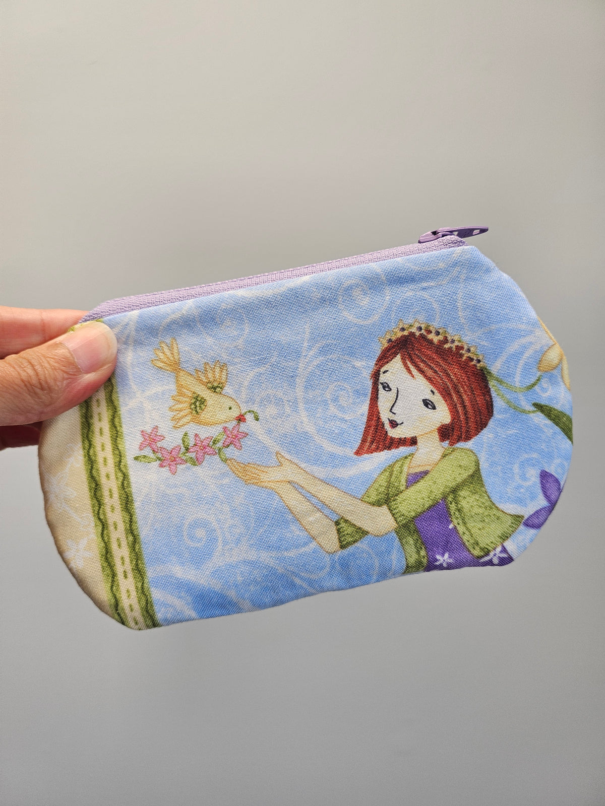 A fabric zippered pouch held by a hand, featuring a whimsical illustration of a girl with red hair, a bird, and flowers on a light blue background.