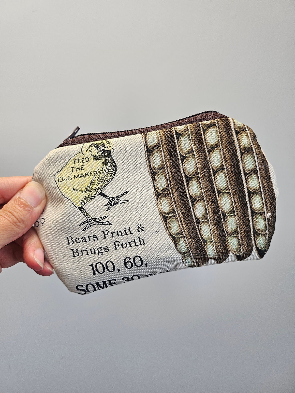 A hand holding a small zippered pouch with a vintage-style egg carton print. The pouch features a cream background with brown egg carton rows, black text reading 'Bears Fruit & Brings Forth 100, 60, SOME 30', and a yellow chick illustration with the words 'FEED THE EGG MAKER'.