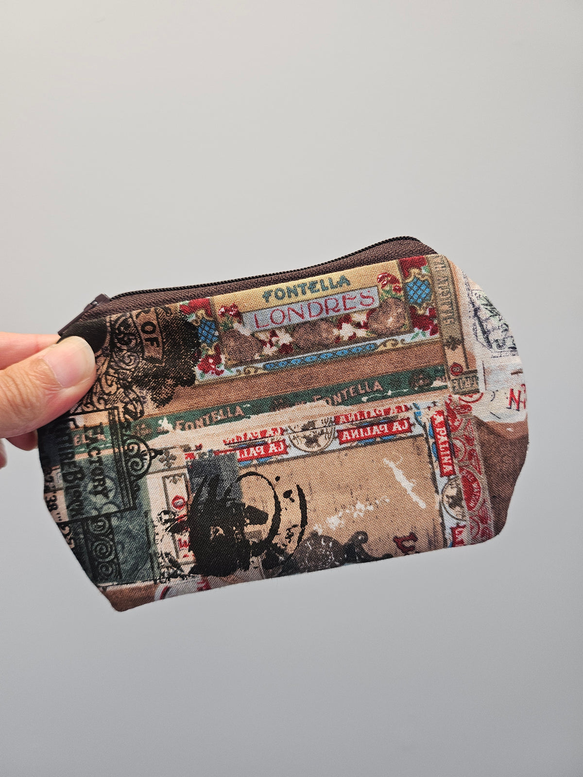 A hand holding a small zippered pouch with a vintage-style collage print featuring postmarks, stamps, and text.
