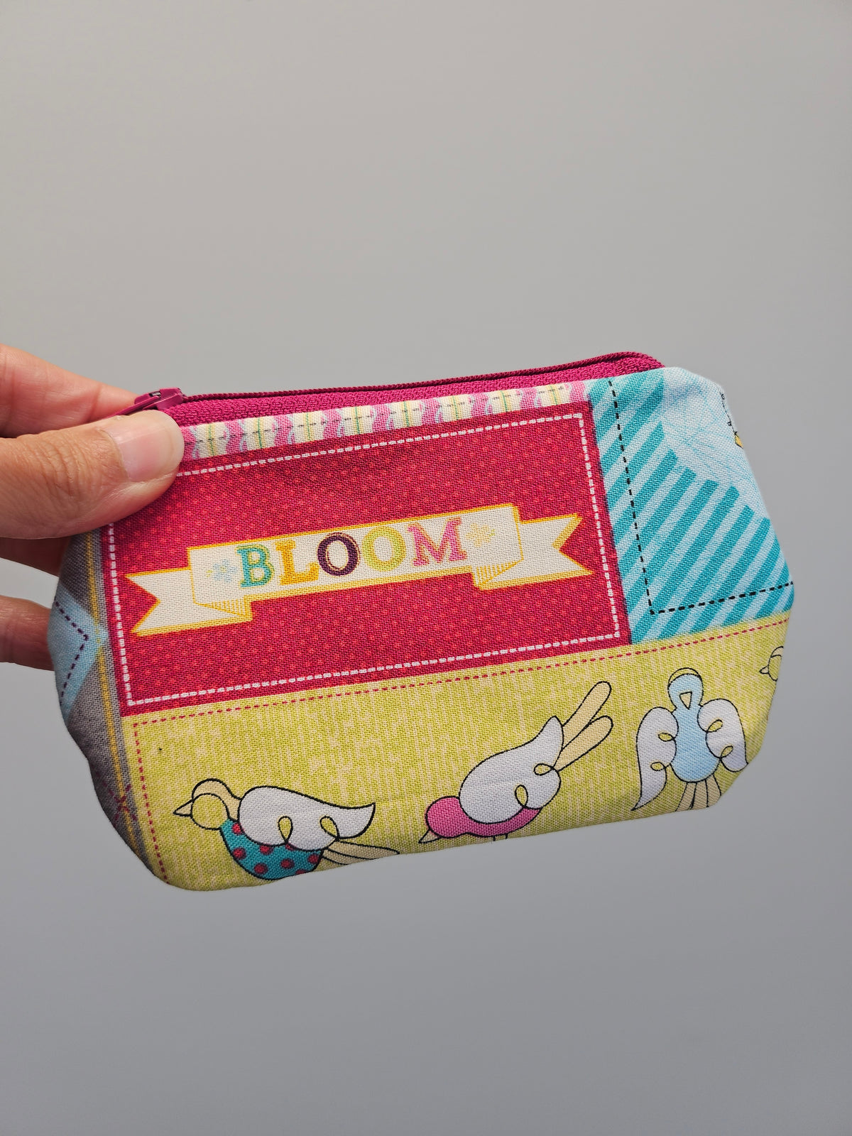 A hand holding a small zippered pouch with a colorful patchwork fabric design featuring the word 'BLOOM' on a red panel and cartoon birds on a yellow panel, against a plain gray background.