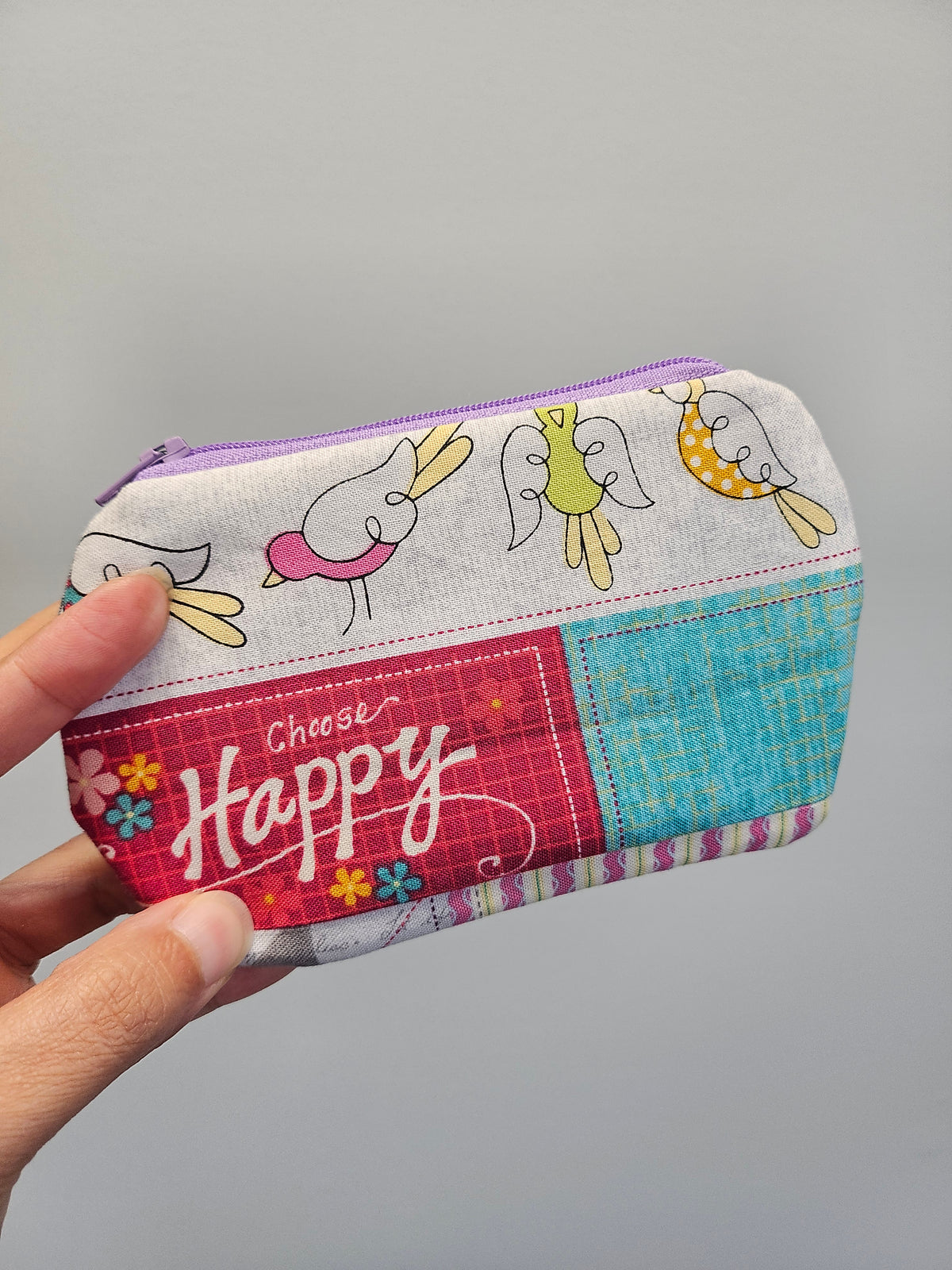 A hand holding a small zippered fabric pouch with a patchwork design featuring cartoon birds, a pink panel with the words 'Choose Happy,' and a turquoise textured section, against a plain light gray background.