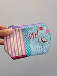 Load image into Gallery viewer, A hand holding a small zippered fabric pouch with a patchwork design. The pouch features pink and cream vertical stripes on the left, a light blue background with diagonal teal stripes, red 'LOVE' text, a small bird in a hot air balloon, and pink and white flowers.
