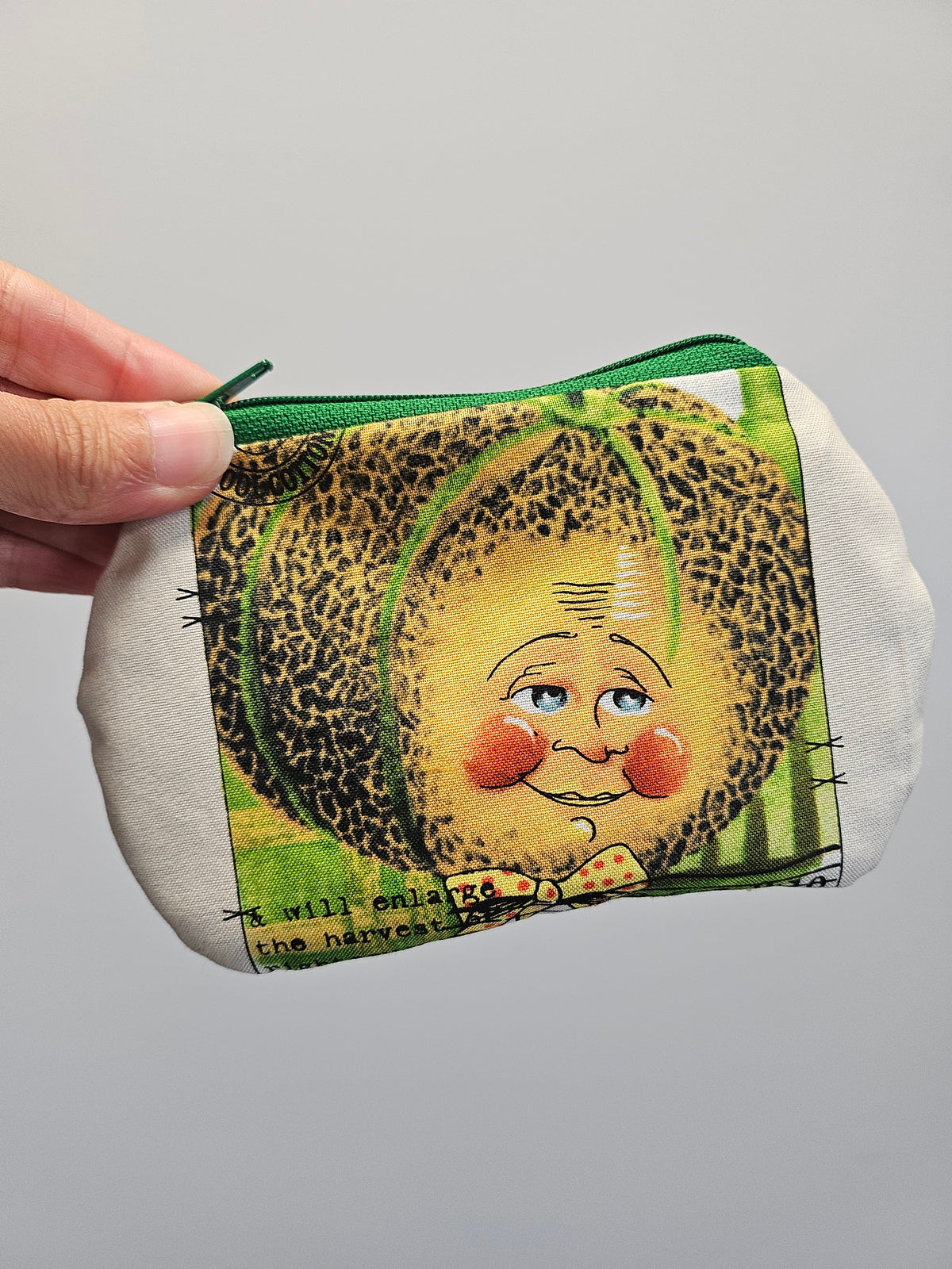 A hand holding a small zippered pouch with a whimsical illustration of a smiling face inside a leopard-print shell, on a plain light gray background.