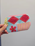 Load image into Gallery viewer, A hand holding a small zippered pouch with a colorful patchwork design featuring red diamonds, pink and yellow stripes, a red flower, and a light blue background.
