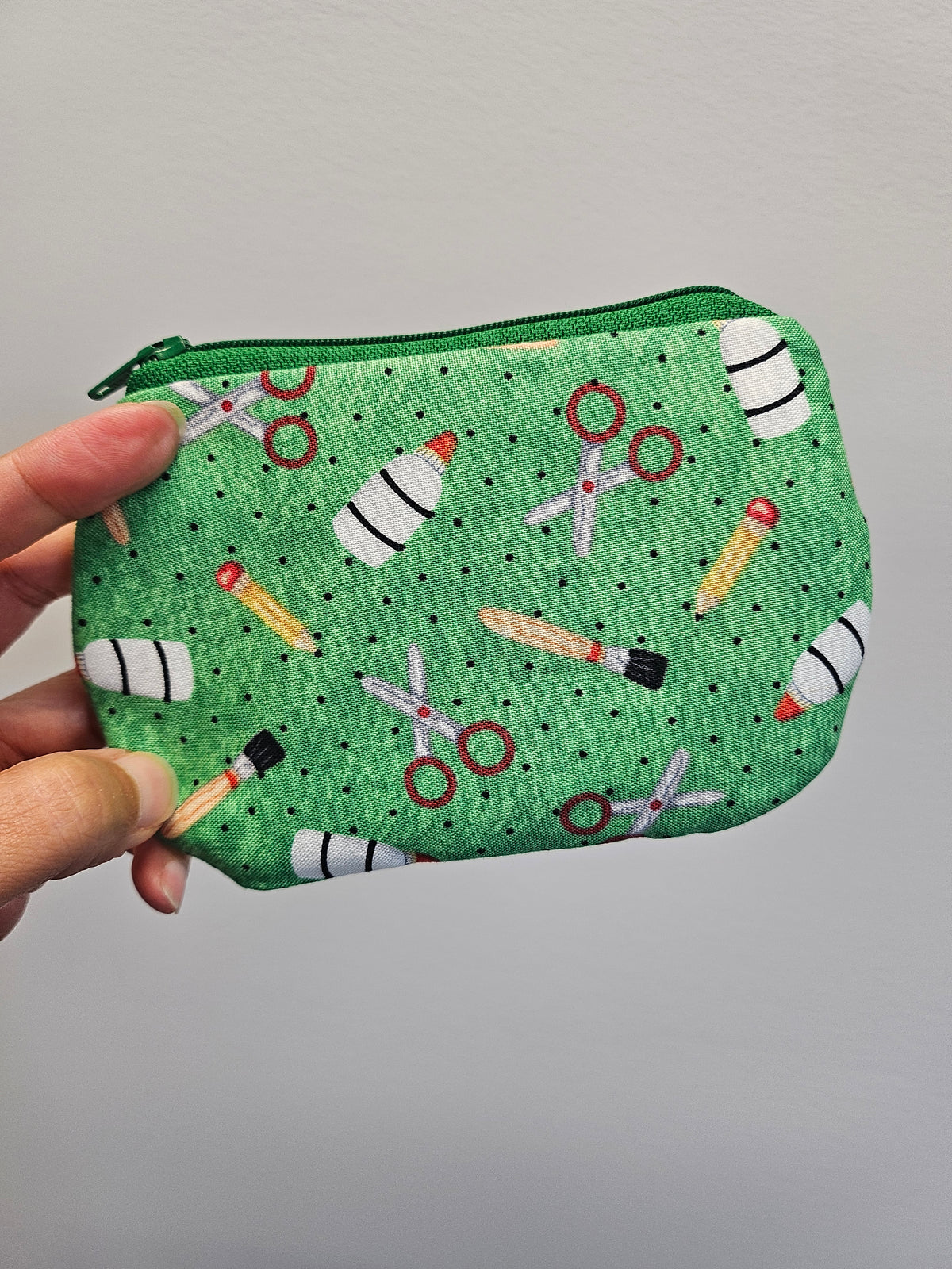 Hand holding a green zipper pouch with school supplies on it against a light gray wall.