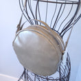 Load image into Gallery viewer, Taupe round crossbody purse hanging on metal dress form.
